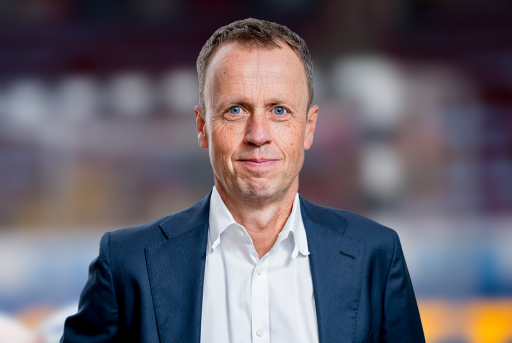 Frank Bohmann, Managing Director of Handball Bundesliga GmbH: Porträt eines Mannes mit kurzen Haaren und blauen Augen, der einen dunkelblauen Anzug und ein weisses Hemd trägt, vor einem verschwommenen Hintergrund.